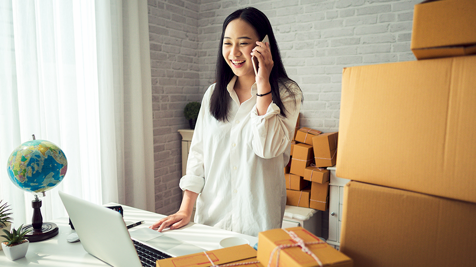Smiling woman on her phone, standing at desk with boxes around her.