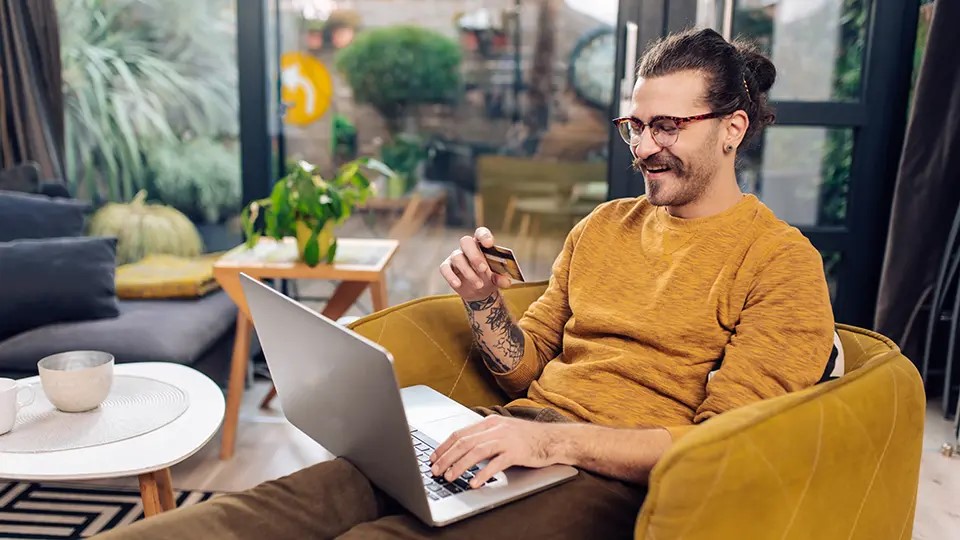 Man on his laptop, looking at his credit card to make a purchase.