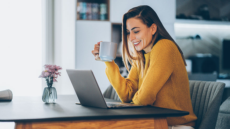 Kvinde sidder og ser på bærbar computer, mens hun holder et kaffekrus i hånden.